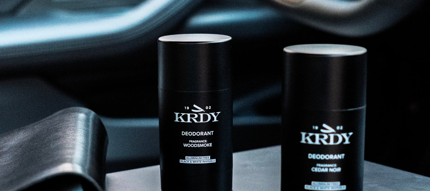 Two KRDY deodorant sticks on a car dashboard with a smartphone and steering wheel in the background.