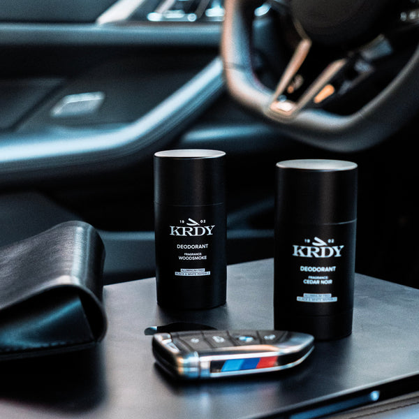 Two KRDY deodorant sticks on a car dashboard with a smartphone and steering wheel in the background.